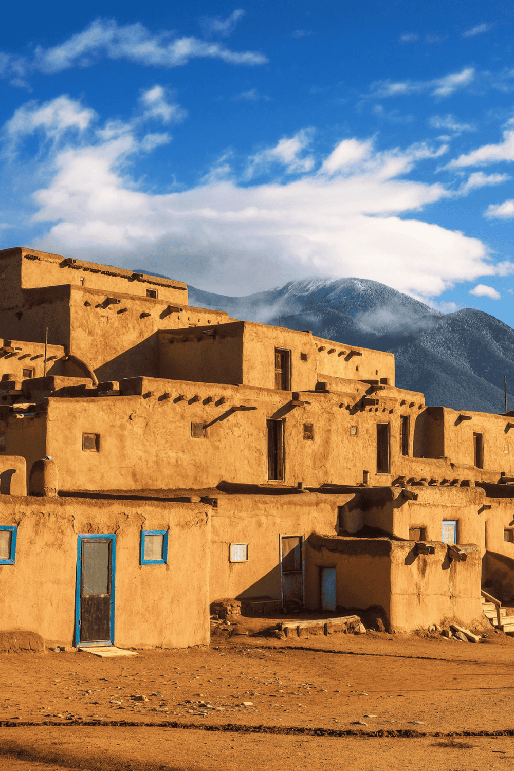 Rustic adobe buildings in New Mexico with mountain backdrop and blue sky, perfect for travel and adventure.