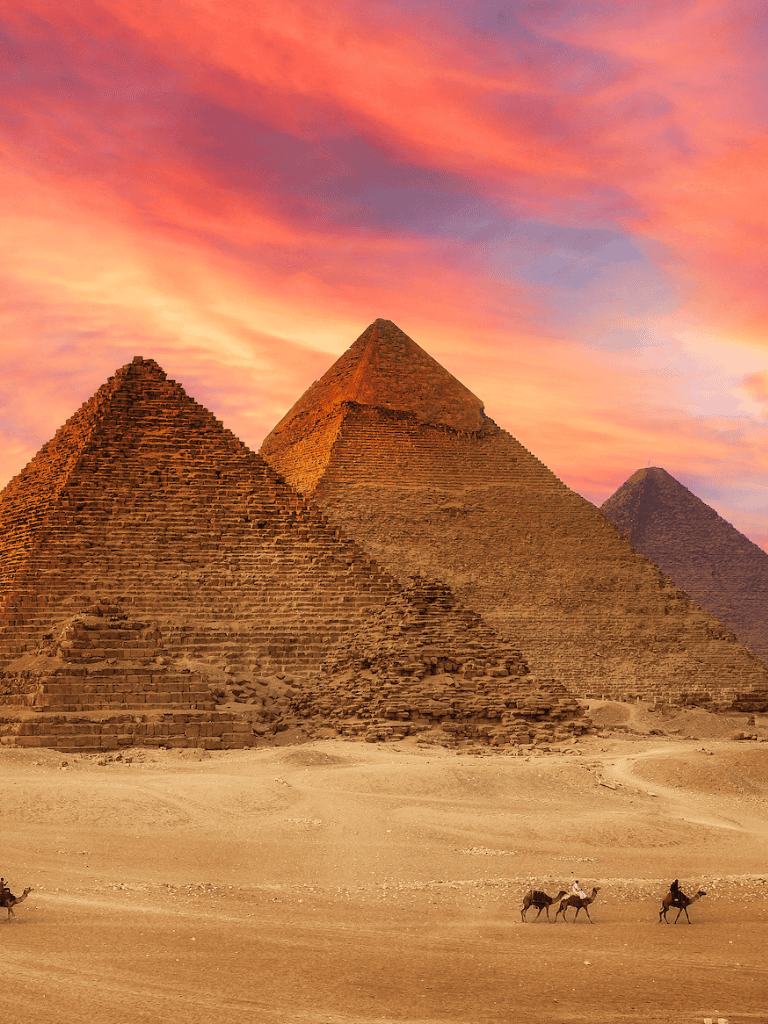 Ancient Egyptian pyramids at sunset beneath colorful sky, desert with camels, historical landmarks, travel destination.