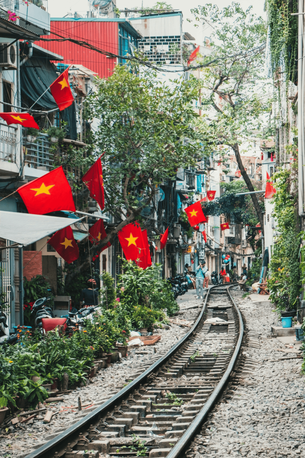 Bright street scene with railway tracks, colorful buildings, green trees, and Vietnamese flags, showcasing vibrant urban life.
