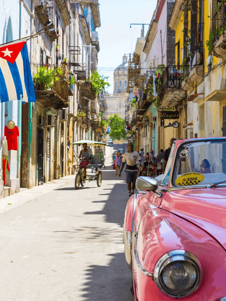 Vintage pink taxi on a colorful street in Havana, Cuba, with vibrant colonial buildings and lively local scene.