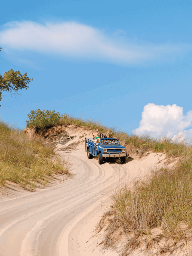 Off-road blue truck driving on sandy trail through green hills under blue sky with clouds.