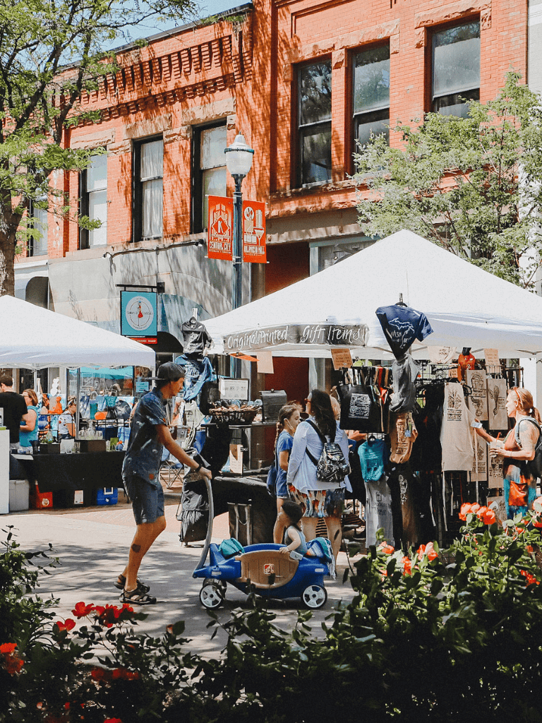 Vibrant outdoor market scene with shoppers, vendor booths, and colorful storefronts in a lively town setting.