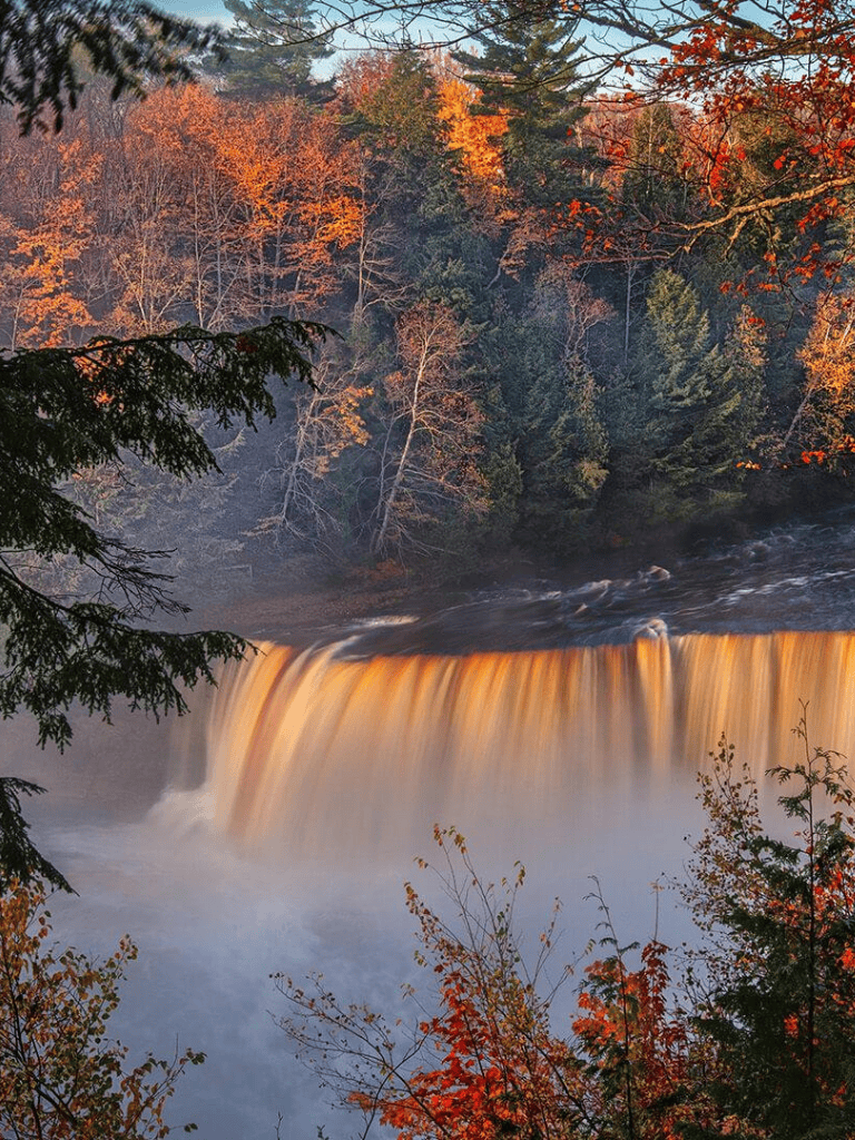 Vivid autumn foliage surrounding a waterfall with mist and flowing water in a scenic forest landscape.