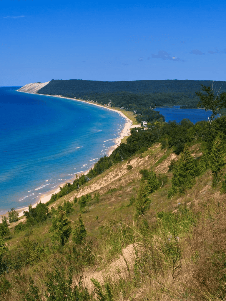 Vast coastal landscape with sandy beach and lush green hills under clear blue sky.