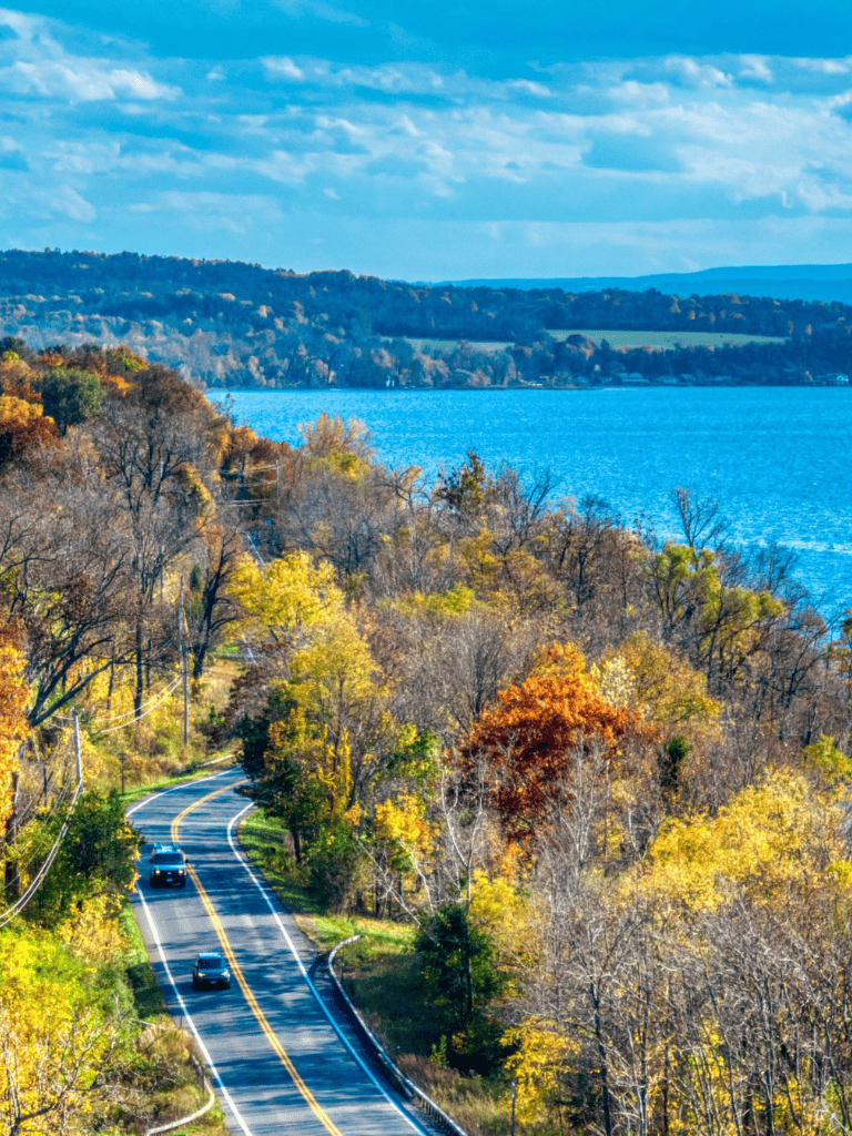 Scenic road trip through autumn foliage near a large lake with colorful trees and rolling hills.