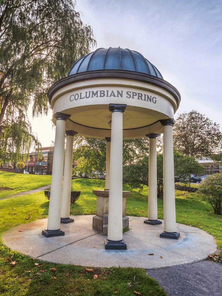 Stone bandstand with "Columbian Spring" sign, surrounded by greenery in Columbia Spring park, inviting visitors to explore.