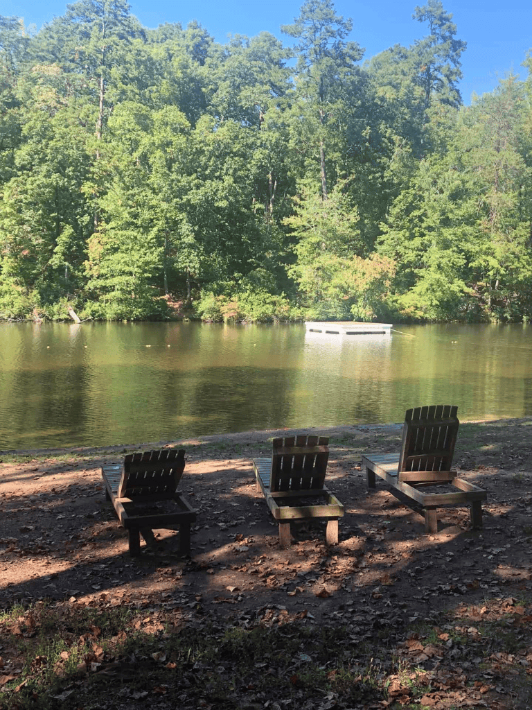 Tranquil lakeside scene with three wooden chairs overlooking the water and lush green trees in the background.
