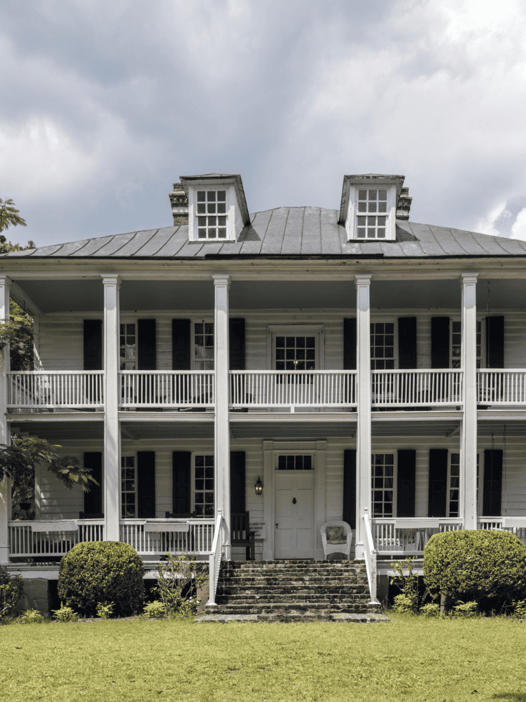 Historic house with white wooden exterior and large porches, located in a scenic outdoor setting.