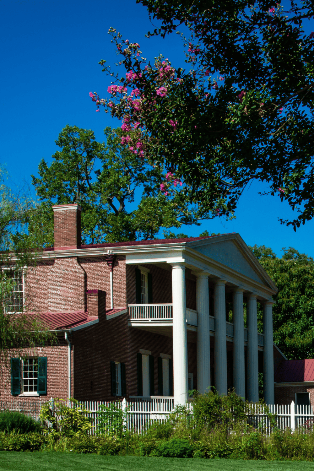 Victorian house with classic columns, lush greenery, and blooming trees under a clear blue sky.