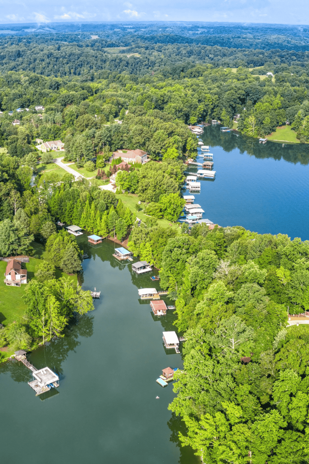 Aerial view of lakeside homes and boat docks surrounded by lush green trees and rolling hills in a scenic outdoor setting.