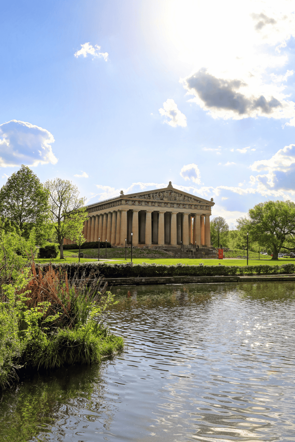 Ancient Greek Parthenon replica near a pond with lush greenery on a sunny day, outdoor historic architecture.