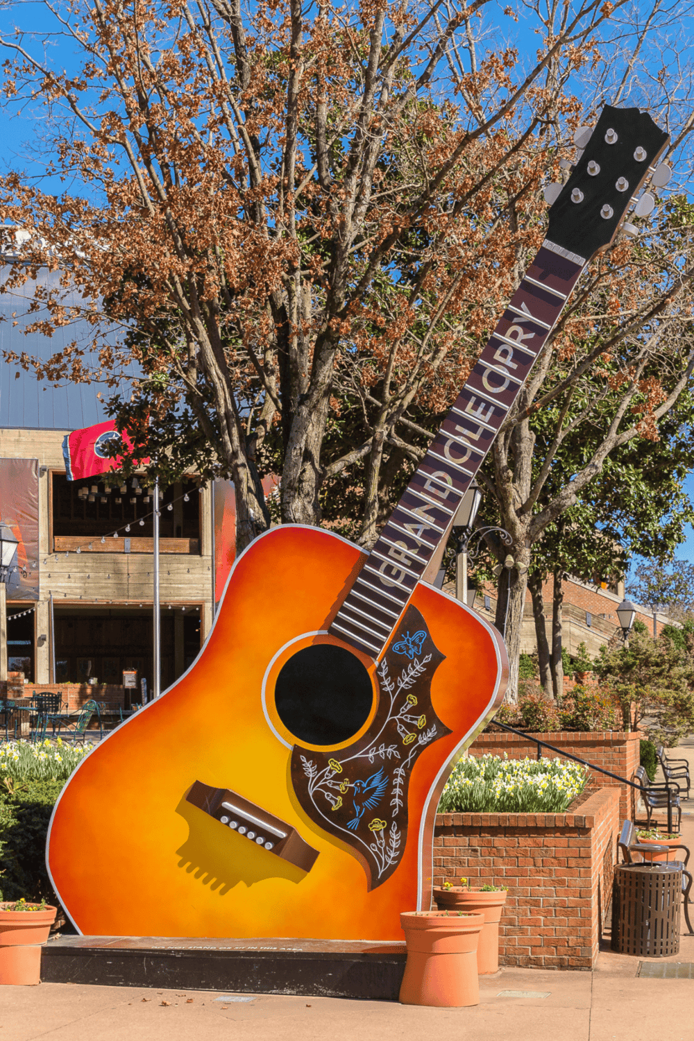 Guitar-shaped outdoor art installation in downtown with colorful floral decoration and trees, perfect for music and art lovers.