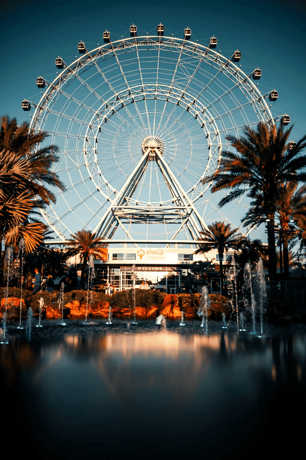 Aerial view of a large Ferris wheel at sunset, with palm trees and water fountains in the foreground.