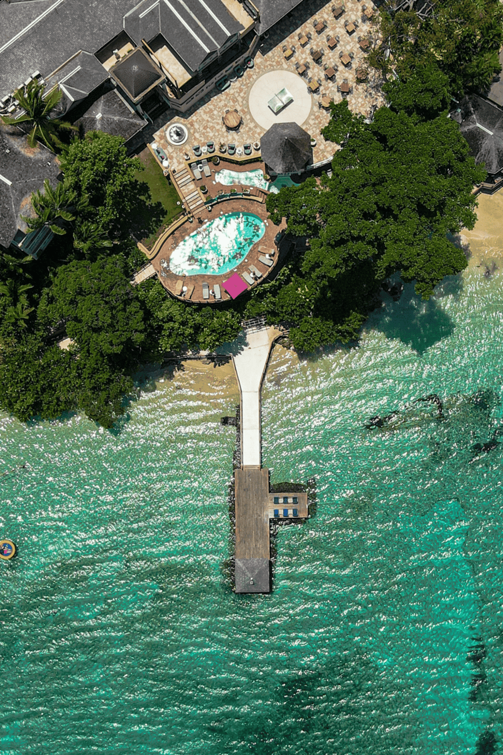 Aerial view of a beachfront resort with a swimming pool, lush greenery, and a pier over clear turquoise water.