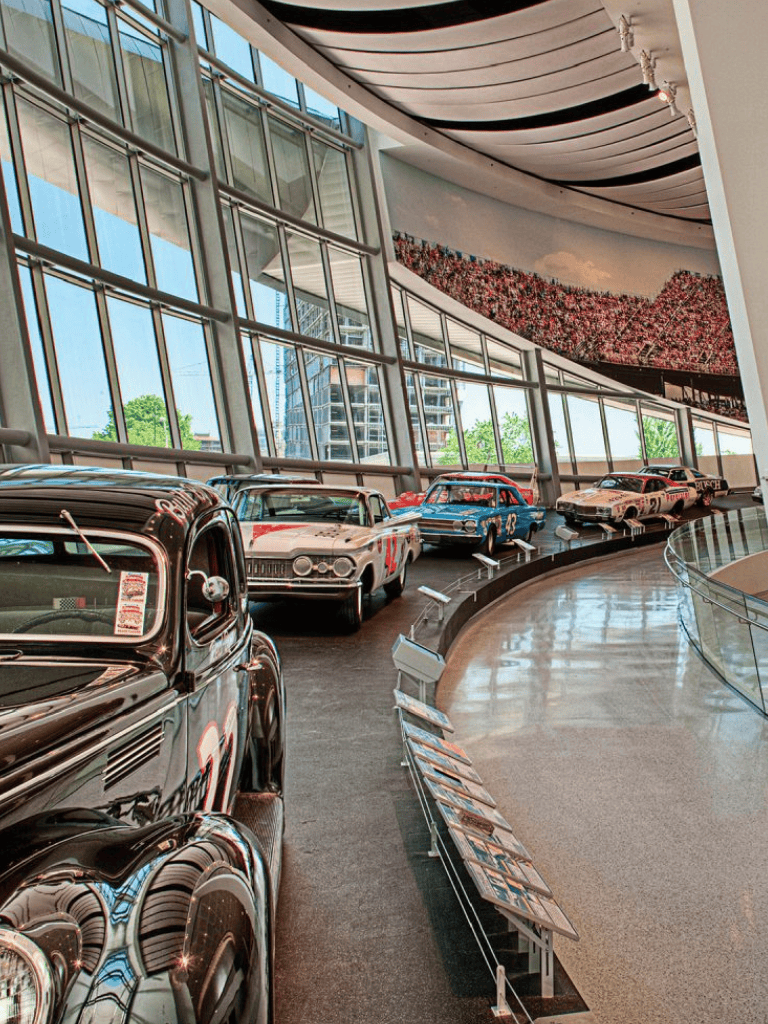Vintage race cars displayed in a modern museum with large windows and artistic mural.