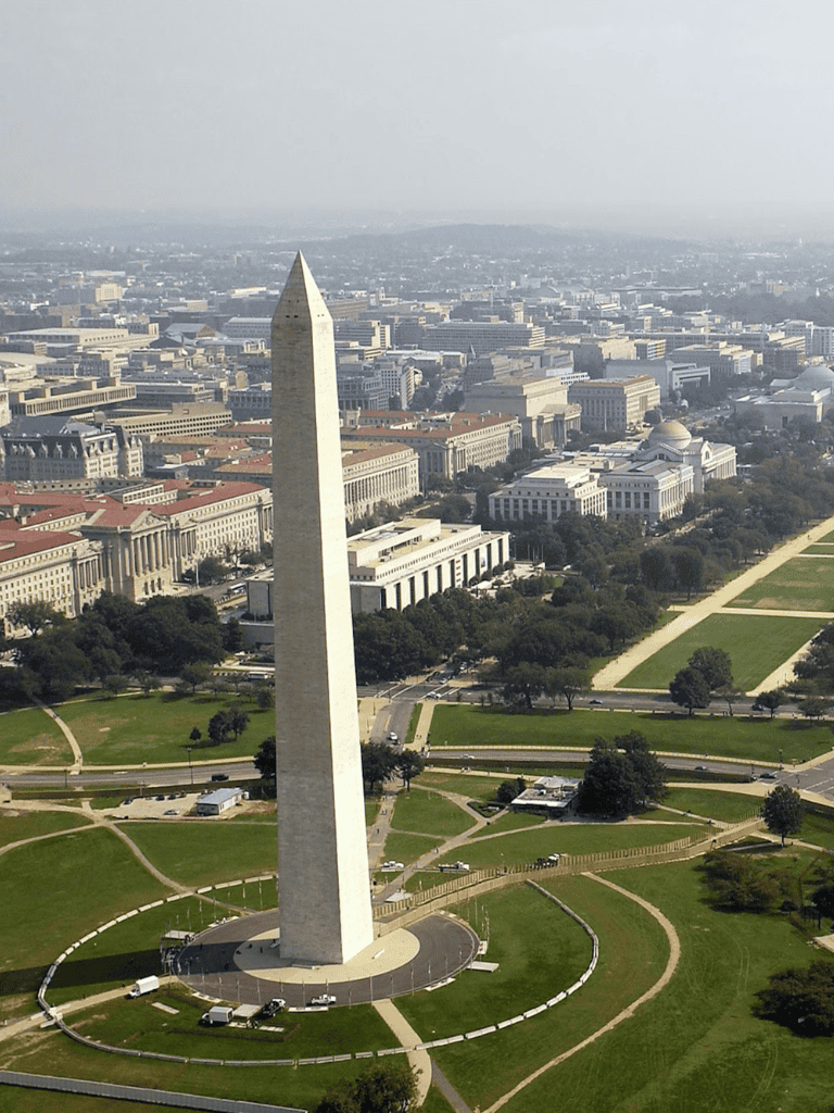 Looming white obelisk in Washington DC atop grassy park with cityscape background.