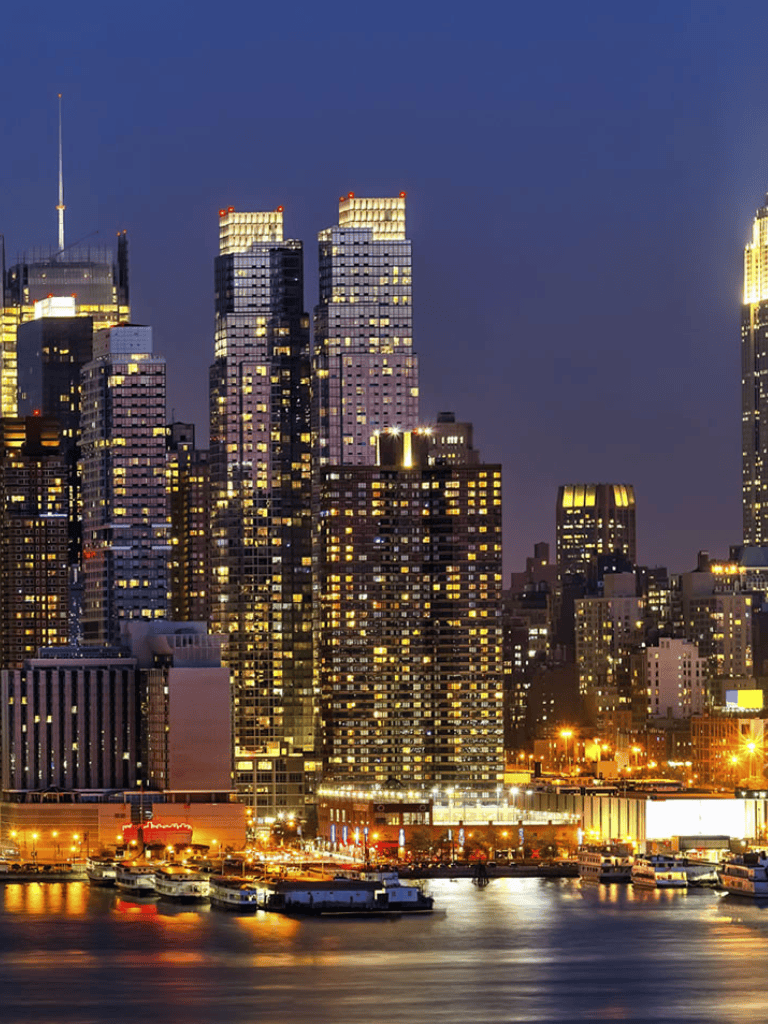 Bright illuminated skyscrapers at night in New York City skyline, waterfront view with boats docked.