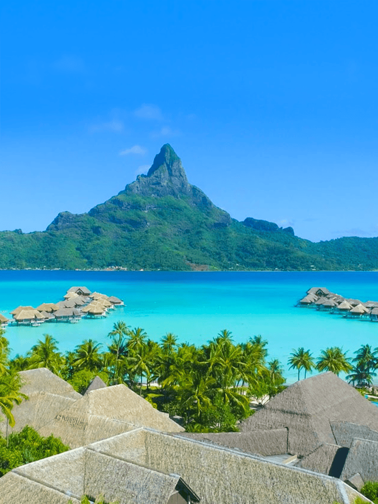 Stunning tropical island view featuring overwater bungalows, turquoise waters, lush greenery, and a mountain backdrop.