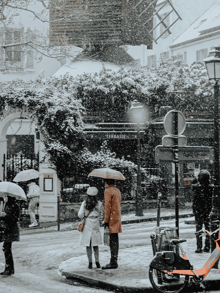 Snowy city street scene with people walking under umbrellas in winter weather.