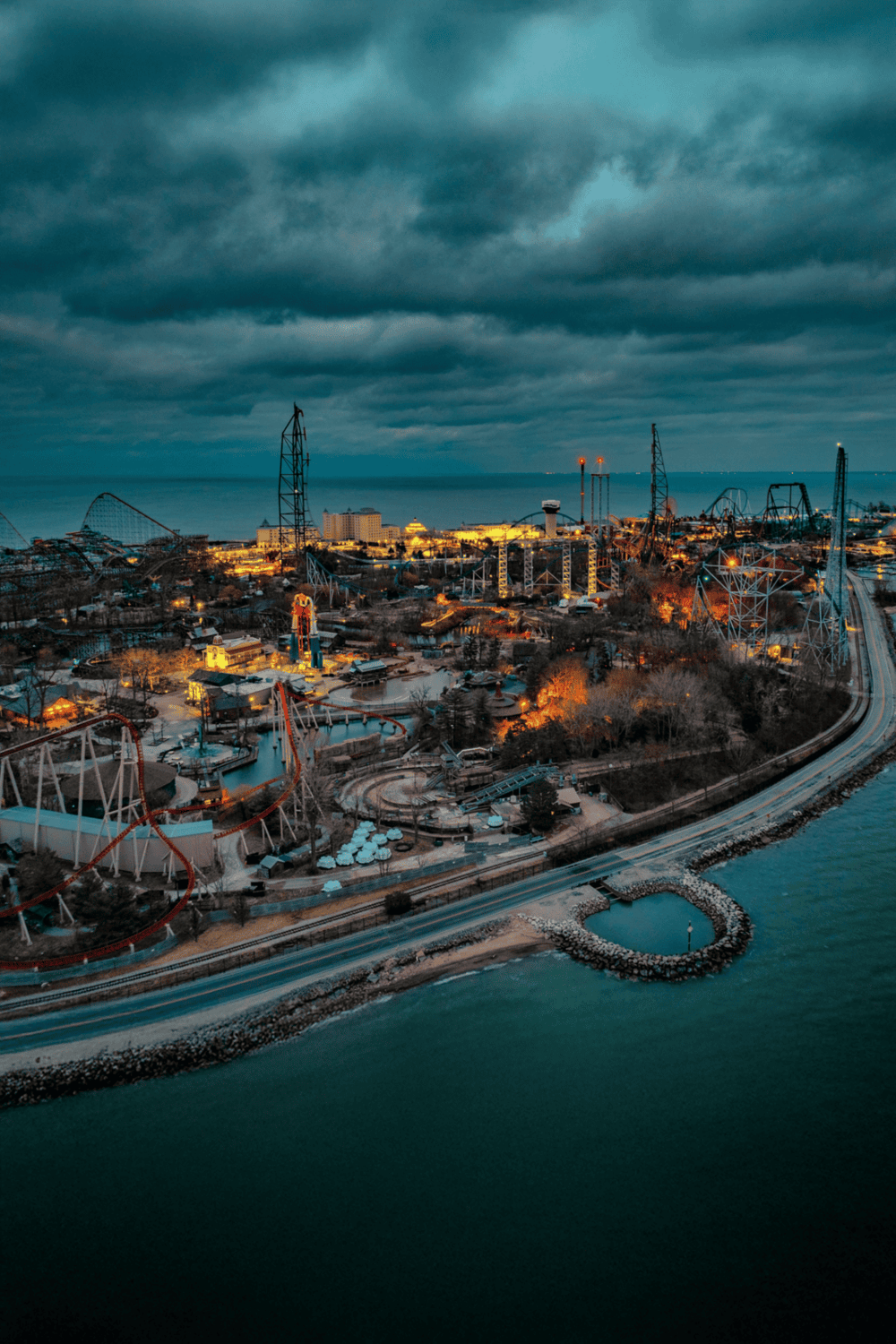 Thrilling amusement park with roller coasters and rides at dusk, near water, under dark stormy clouds.