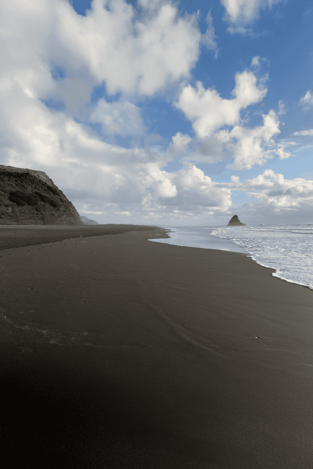 Serene beach landscape with dark sand, cliffs, and a distant rock formation under a partly cloudy sky.