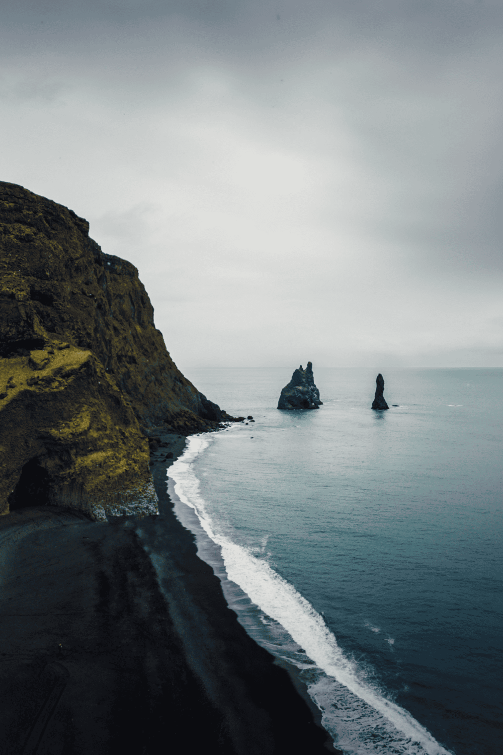 Cliffside coastal landscape with sea stacks, black sand beach, and overcast sky, promoting travel and adventure.