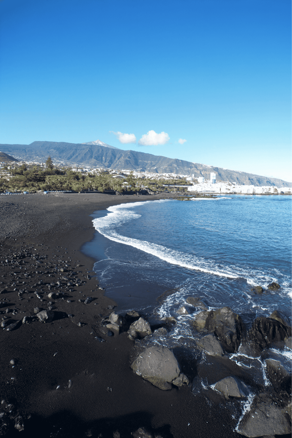Serene black sand beach with mountains in the background, scenic coastal view, and clear blue sky.