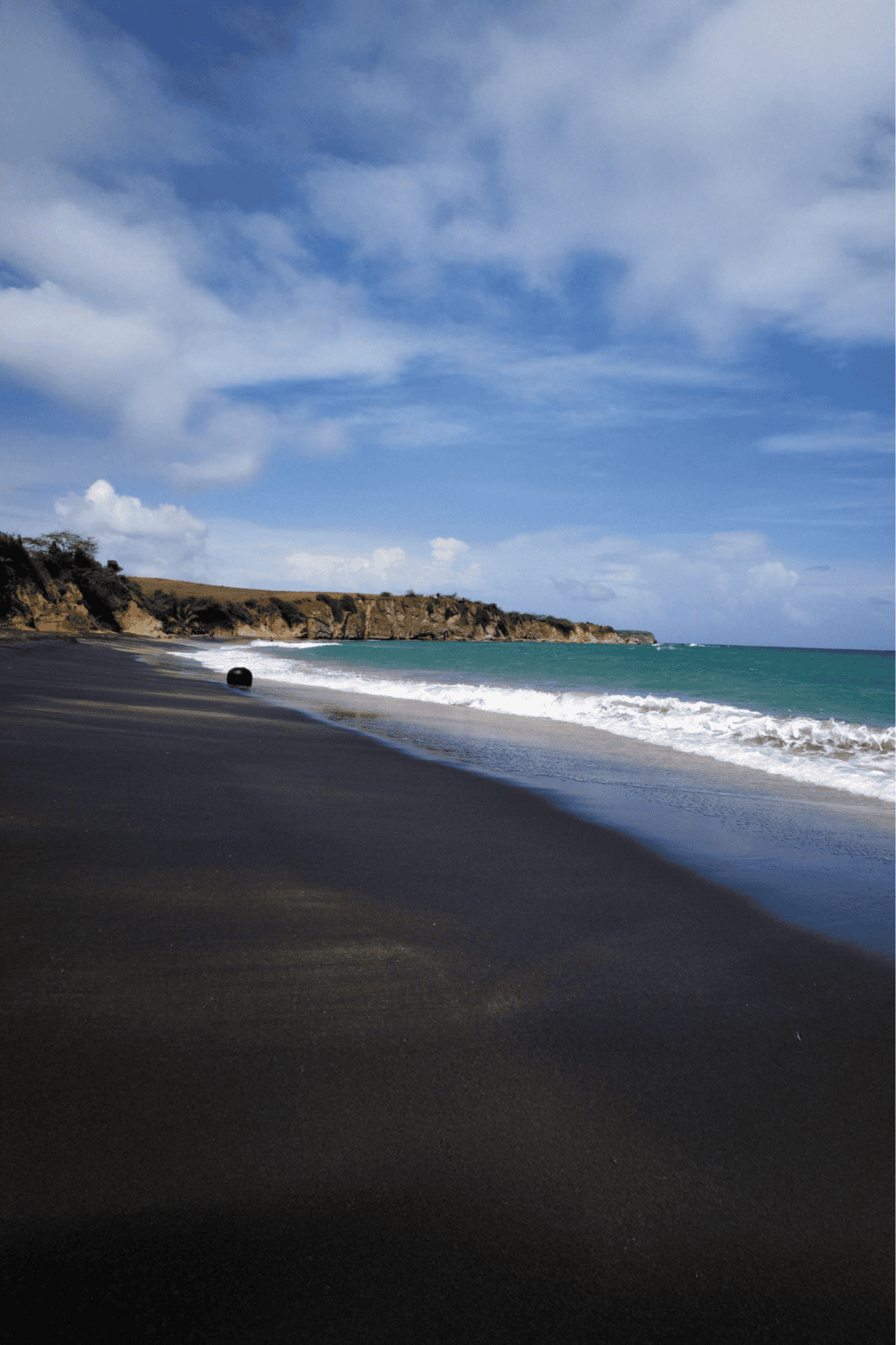 Secluded beach with dark sand, blue ocean, and scenic cliffs under partly cloudy sky.