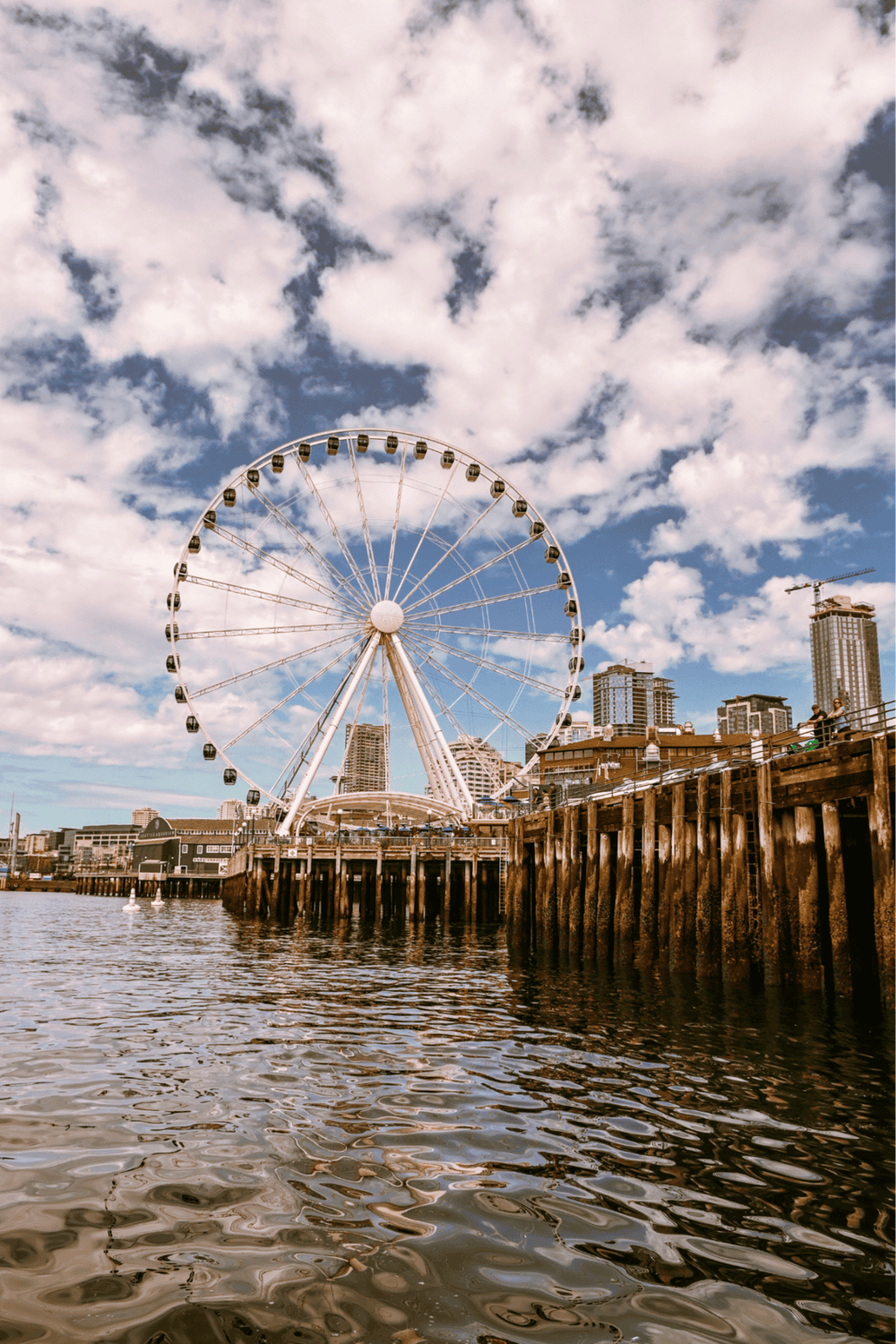 Aerial view of large Ferris wheel on waterfront with city skyline in background.