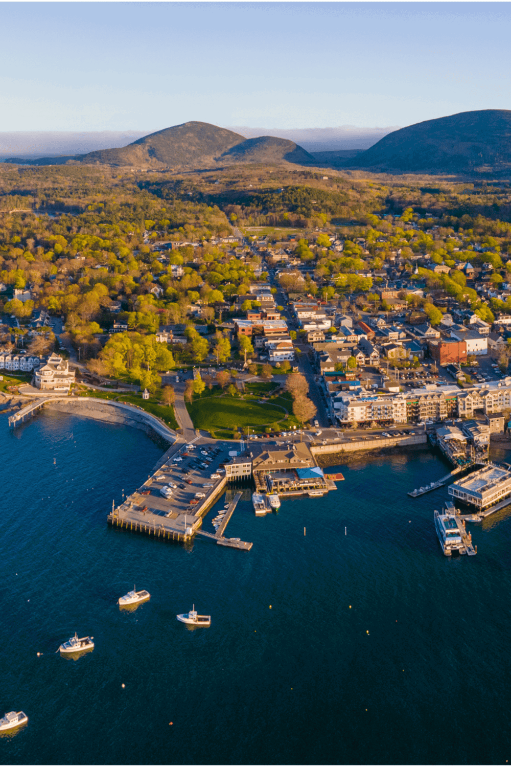 Aerial view of a coastal town with harbor, boats, lush trees, and surrounding mountains, promoting navigation and directional services.