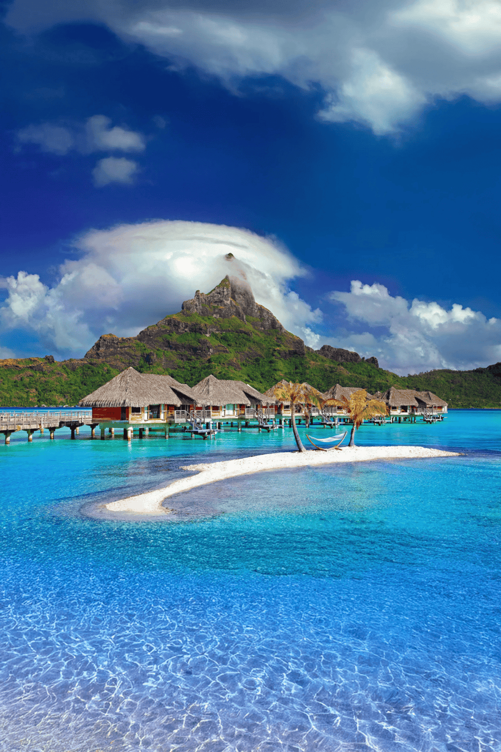 Tropical overwater bungalow resort on turquoise lagoon with lush mountain backdrop and clouded sky.