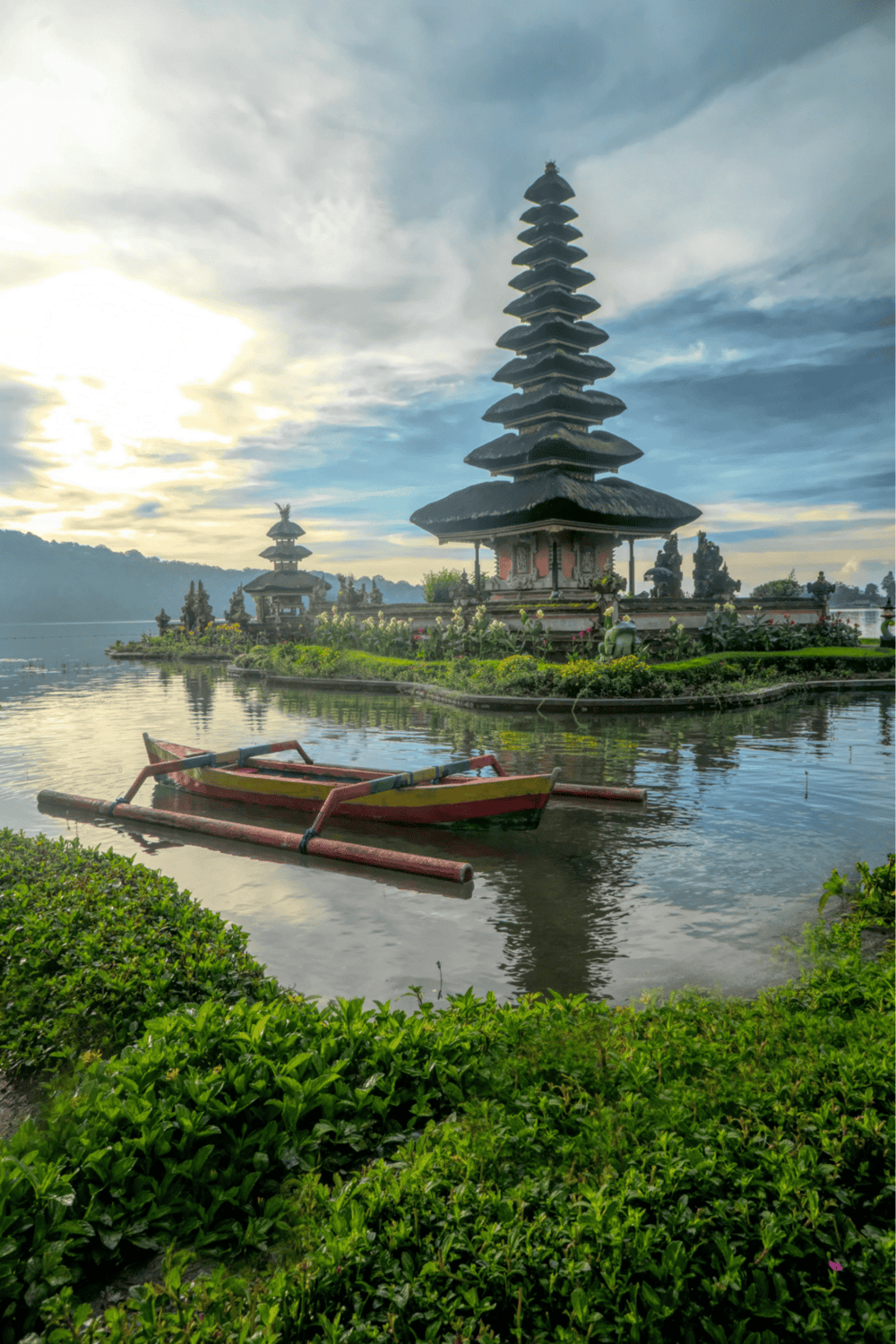 Ancient Balinese temple on a tranquil lake with traditional boat and lush greenery at sunset.