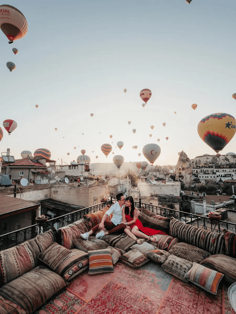 Hot air balloons over Cappadocia in Turkey with a couple relaxing on a cozy rooftop lounge.