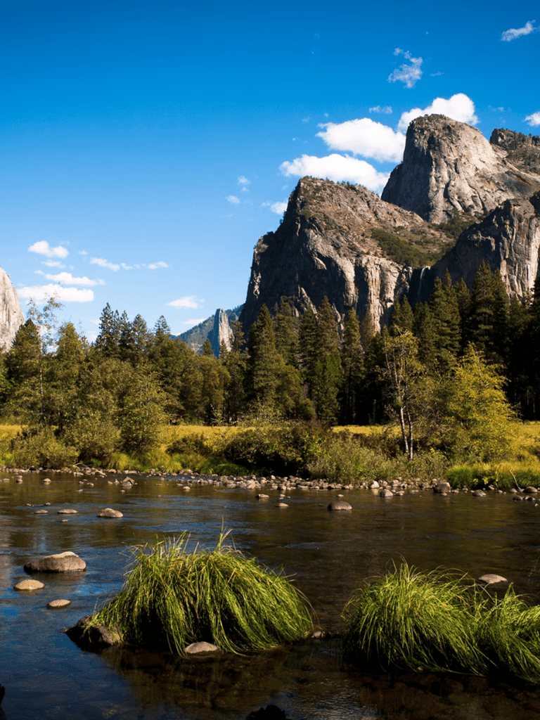 Vast mountain landscape with rocky peaks, lush forest, and a flowing river under a bright blue sky.