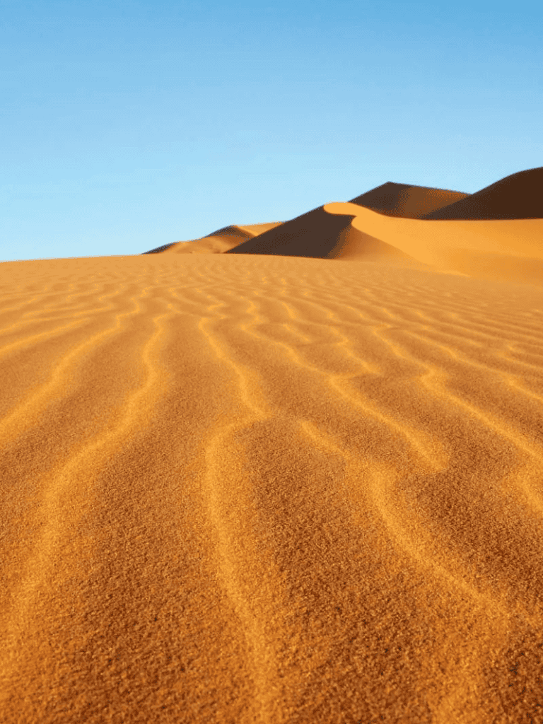 Soft golden desert sand dunes with ripples and clear blue sky, ideal for outdoor adventure and travel exploration.