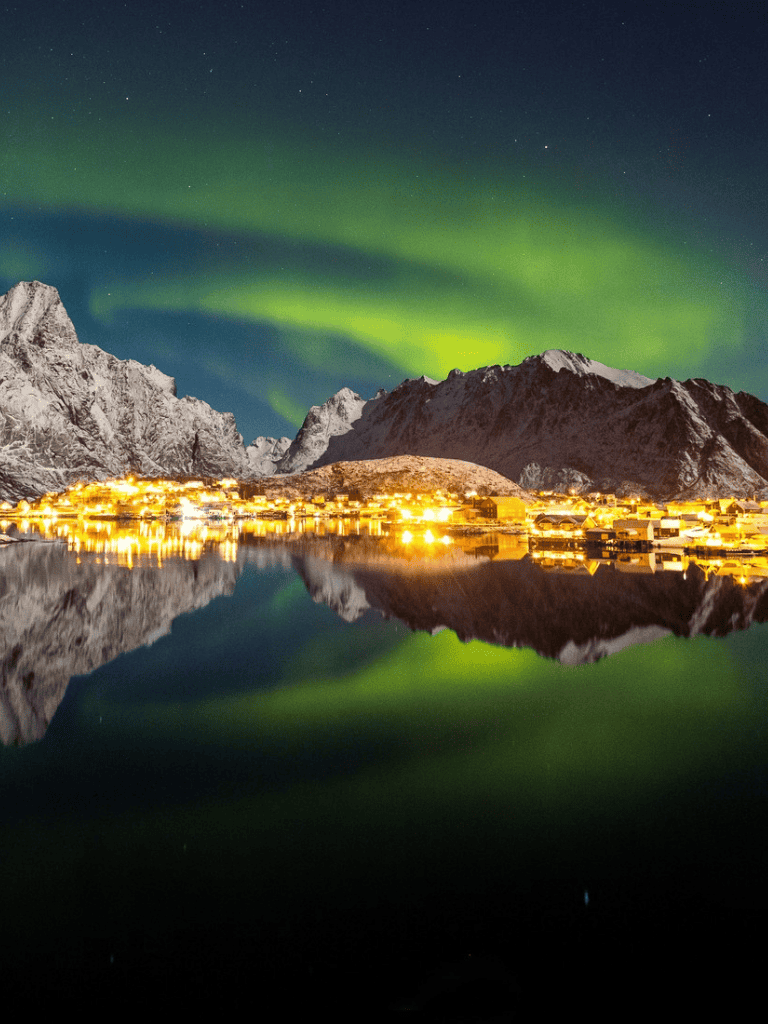 Northern Lights over mountain village reflected in fjord, scenic Norway travel destination.