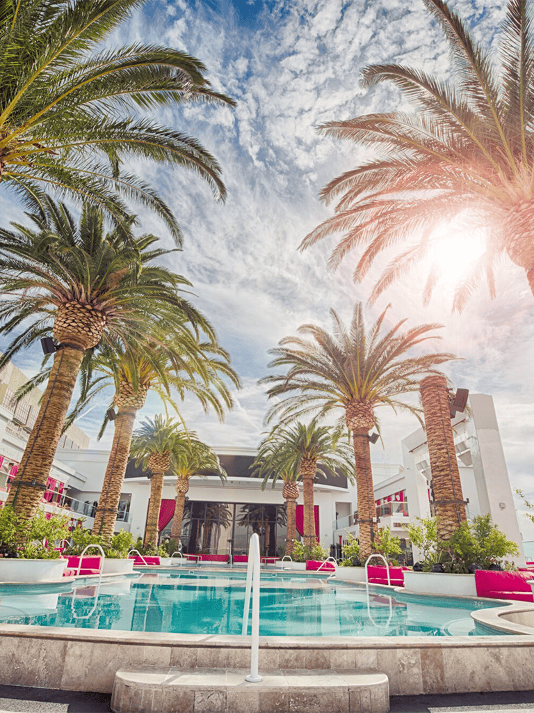 Palm trees near luxury hotel pool with modern architecture and vibrant seating, sunny sky with clouds.