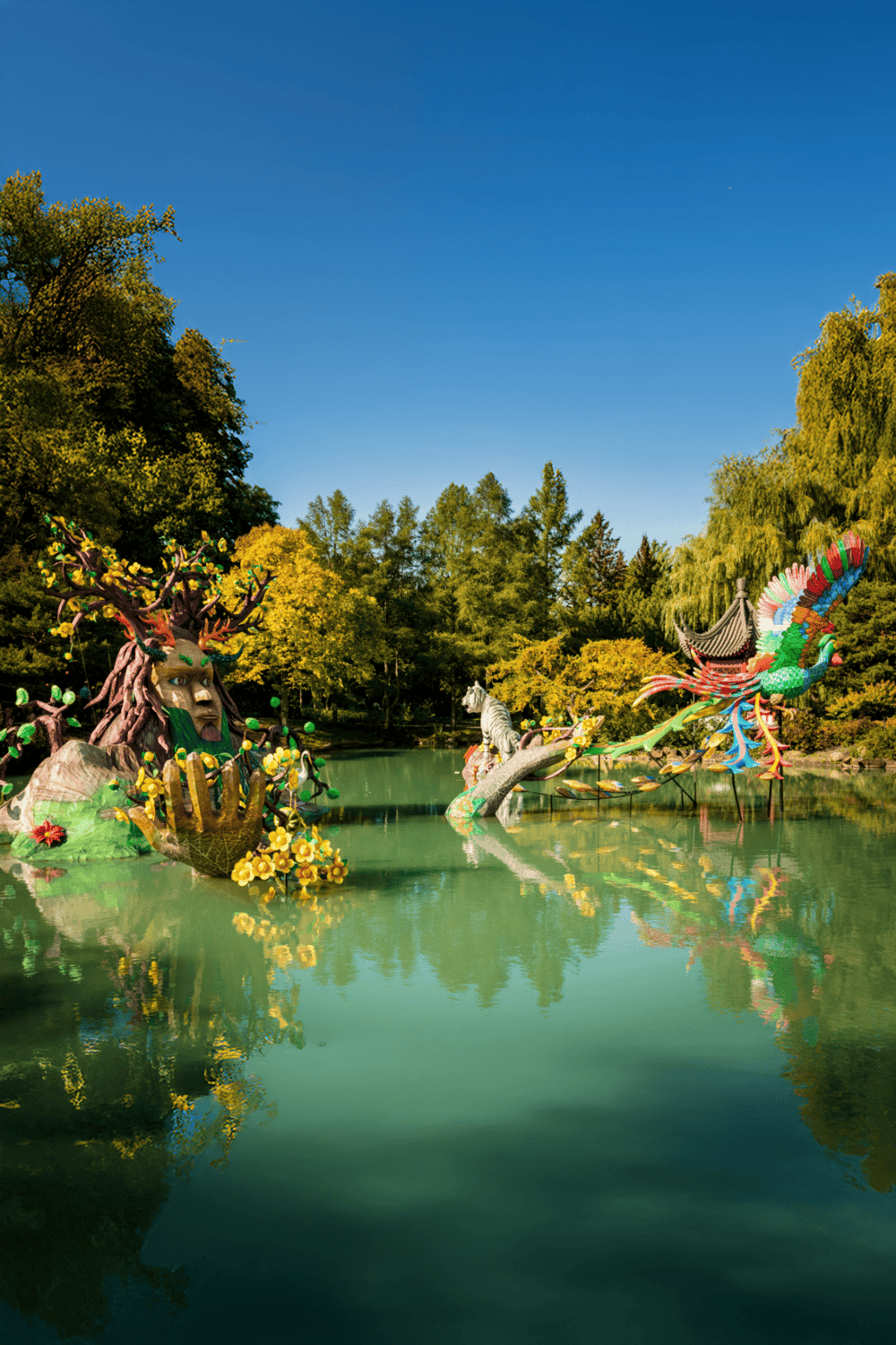 Vibrant colorful sculptures of a dragon, tiger, and phoenix in a scenic park pond, with lush trees and clear blue sky background.