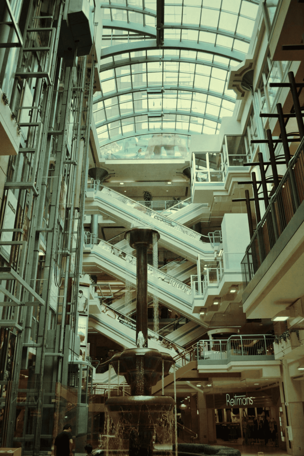Indoor shopping mall with water fountain, escalators, and glass ceiling.