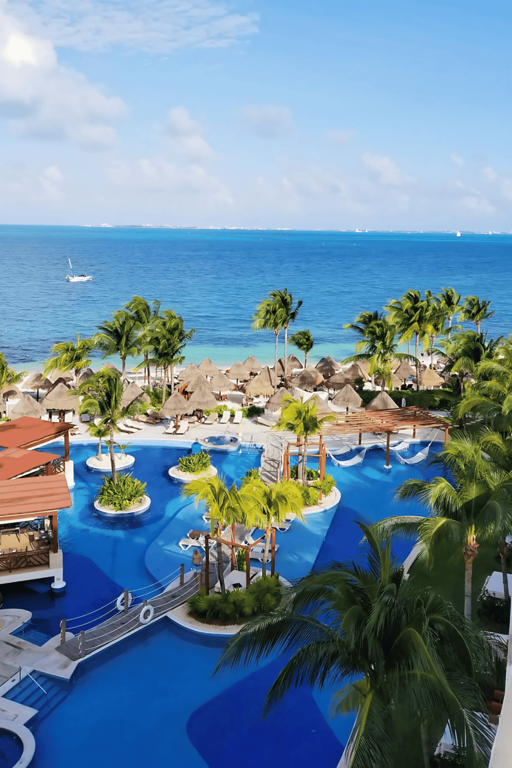 Luxury resort pool overlooking ocean with palm trees and thatched umbrellas in sunny tropical paradise.