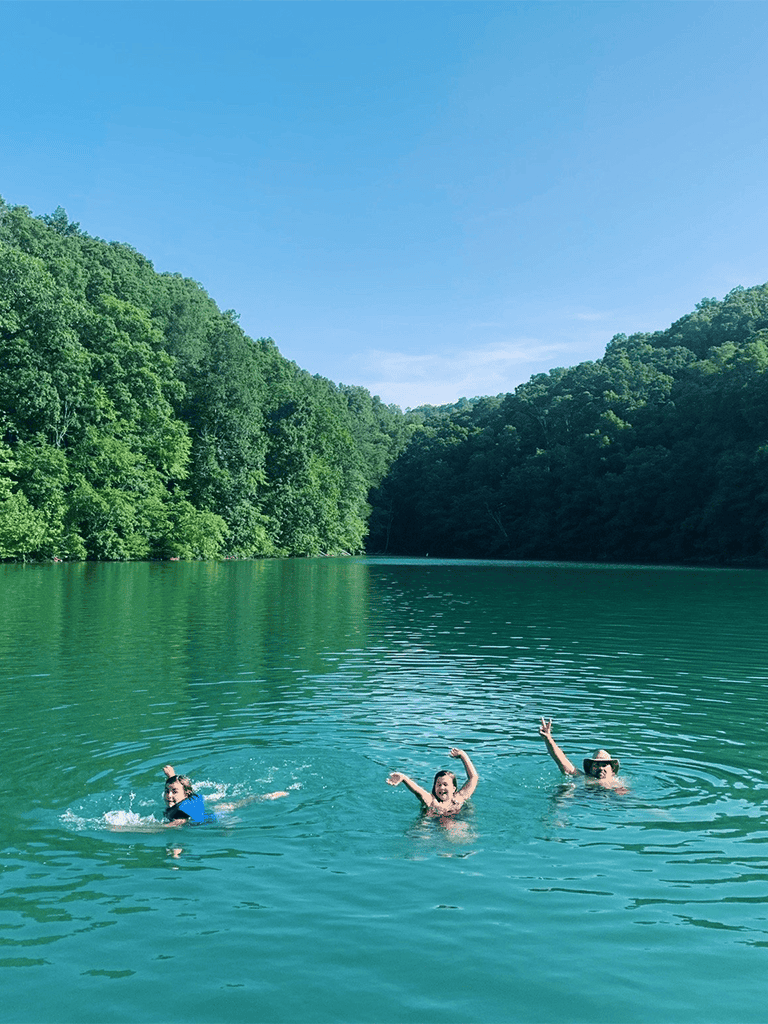Kids swimming in a peaceful lake surrounded by lush green trees and clear blue sky.