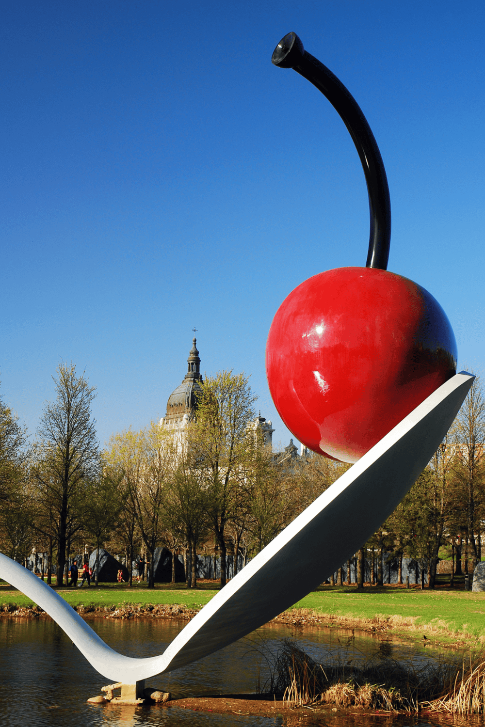 Bright red cherry sculpture in a city park with historic building in background.