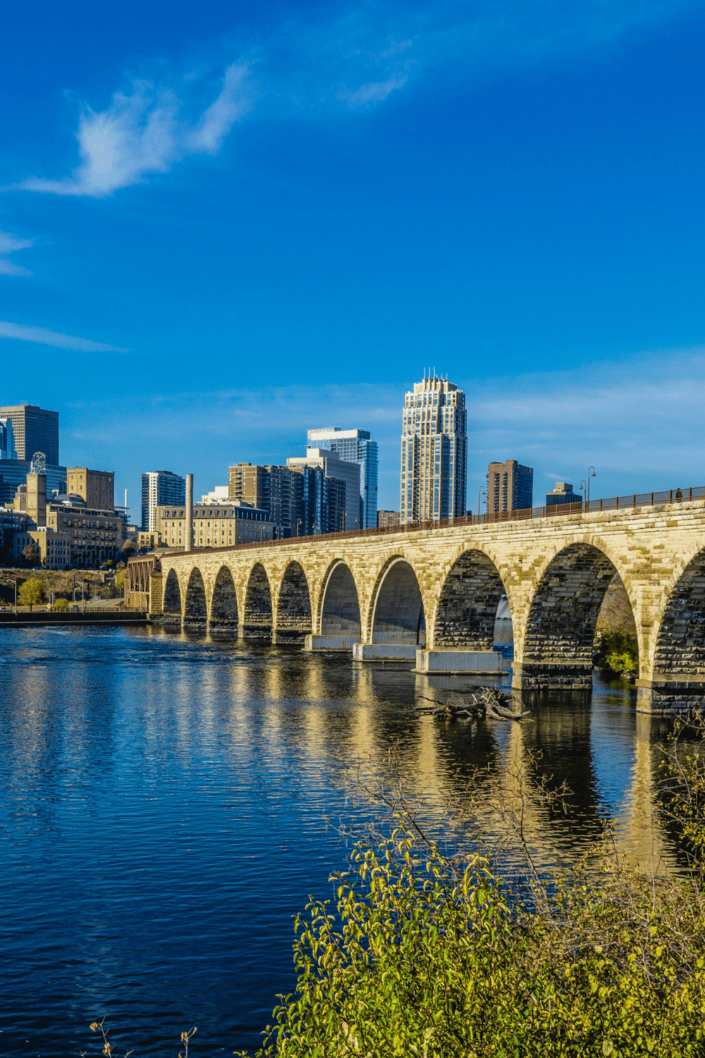 Historic stone bridge over river with city skyline under blue sky, Portland Oregon.