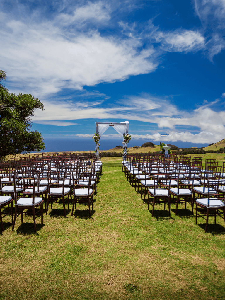 Elegant outdoor wedding setup with chairs and floral arch on scenic coastline background.