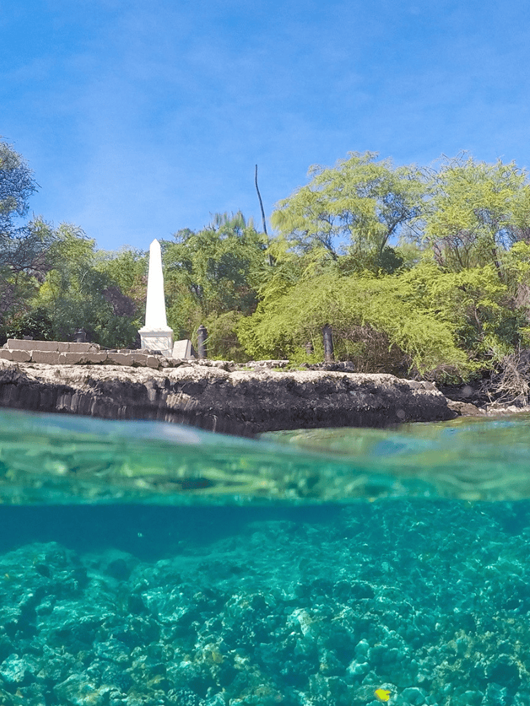 Serene lakeside scene with lush greenery, clear blue sky, and a memorial monument on the shore.