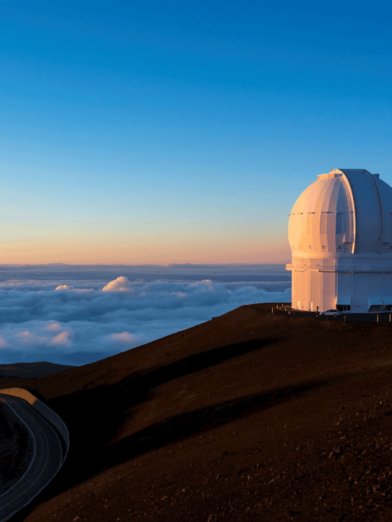 Clear night sky over a large observatory on a mountain peak, overlooking clouds and horizon.