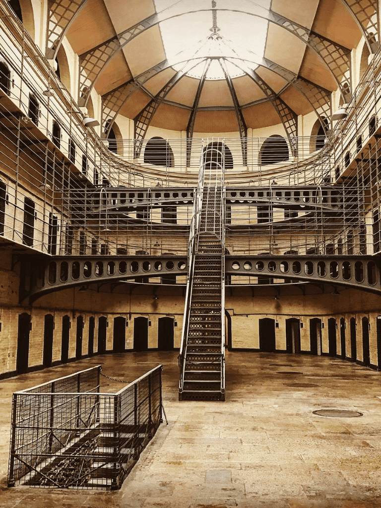 Rustic prison interior with metal walkways and central staircase, historic jail cell blocks, and domed ceiling.