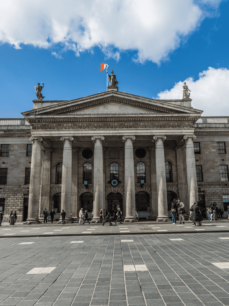 Grand neoclassical building with columns and statues, city street scene with pedestrians.