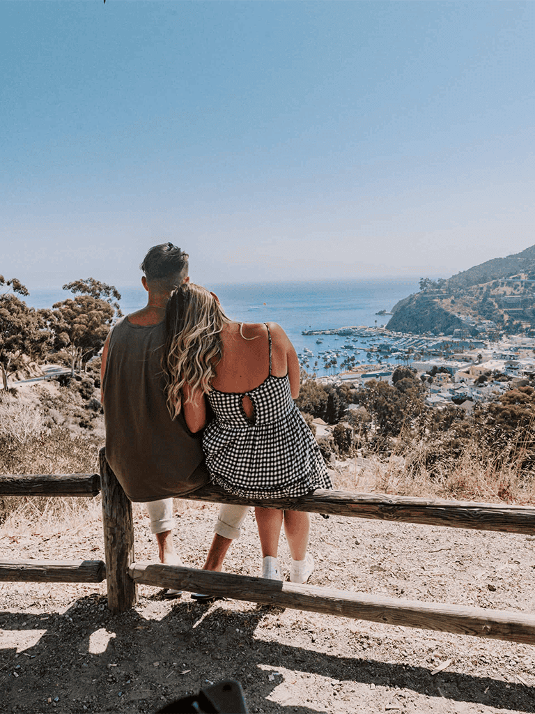 Scenic view overlooking a coastal town and marina with a couple sitting on a wooden fence.