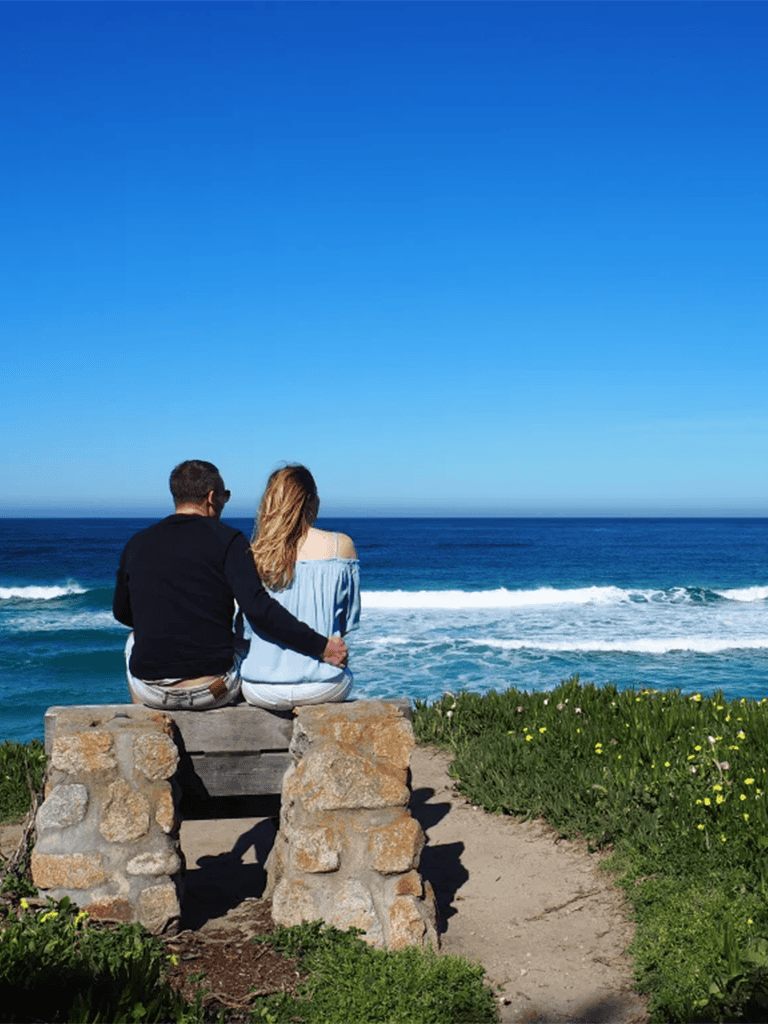 Romantic couple sitting on stone bench overlooking ocean, sunny day, scenic coastal view, travel and relationship moments.